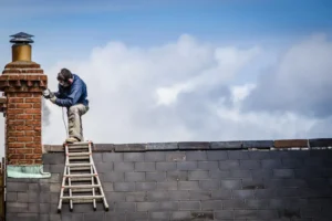 Artisan utilisant une meuleuse d'angle sur une cheminée en briques depuis une échelle posée sur un toit incliné.